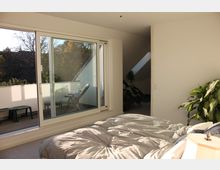 Helles Schlafzimmer mit einem großen Bett, einer Nachttischlampe und Zugang zu einem Balkon mit Schiebetür. Der Balkon ist mit Stühlen und einem kleinen Tisch ausgestattet, im Hintergrund sind Bäume und ein Gebäude zu sehen.