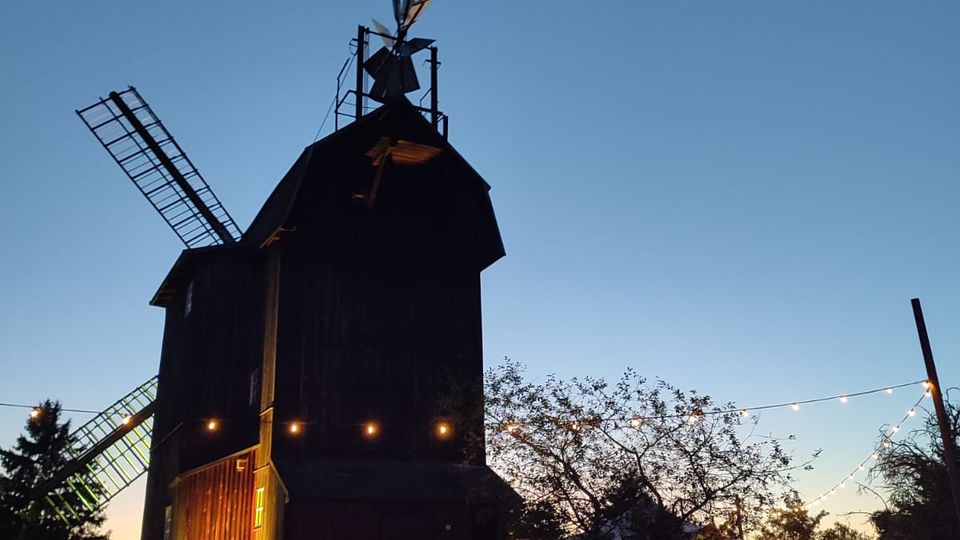 Holzverkleidete Windmühle mit seitlichen Flügeln und kleinem Aufbau an der Spitze, aufgenommen in der Dämmerung vor blauem Himmel. Im Vordergrund eine Wiese mit Lichterketten zwischen Bäumen und einem überdachten Sitzbereich.