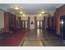 Großer Eingangsbereich mit hohen Decken, Wänden aus Holzvertäfelung und einem glänzenden Steinboden. Eine rot ausgerollte Teppichbahn führt zur Mitte des Raumes, wo eine Büste an der Wand hängt; an den Seiten befinden sich Türen mit Glaselementen.