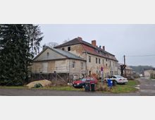 Großes, teilweise renovierungsbedürftiges Gebäude aus hellem Mauerwerk mit Ziegeldach; links ein einstöckiger Anbau mit Gerüst; davor parkende Autos, Mülltonnen und ein Verkehrsschild; Umgebung mit Bäumen, Grasflächen und unbefestigten Wegen.