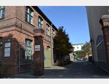 Backsteingebäude in einem Hof mit schmiedeeisernem Tor, hohen Fenstern und grünen Türen; rechts ein gepflasterter Weg zwischen Gebäuden, Bäume und Fahrräder im Hintergrund.