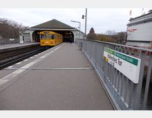 Außenaufnahme der U-Bahn-Station Hallesches Tor in Berlin, mit einem gelben U-Bahn-Zug auf der linken Seite, der in die Station einfährt. Rechts ist das Stationsschild sichtbar und im Hintergrund stehen Bäume, ein Fluss und ein historisches Gebäude mit Kuppel.