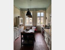 Helle Landhausküche mit hohen Fenstern, die auf eine Backsteinfassade blicken. Die Küche verfügt über weiße Holzschränke, eine zentrale Arbeitsinsel mit dunkler Holzplatte und schwarze Hängelampen über dem Tisch, mit roten Fliesen als Bodenbelag.