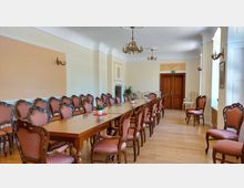 Eleganter Konferenzraum mit langen, hölzernen Tischen und gepolsterten Stühlen mit ornamentierten Holzrahmen. Der Raum hat helle Wände, dekorative Kronleuchter und Wandverzierungen, sowie eine Tür aus Holz und große Fenster, die Tageslicht hineinlassen.