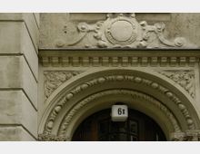 Dekoratives Eingangsportal eines historischen Gebäudes mit einem verzierten Rundbogen, floralen Stuckelementen und einer Hausnummer „61“ über der dunklen Holztür. Die Fassade besteht aus hellem, verputztem Stein mit detaillierten Ornamenten.