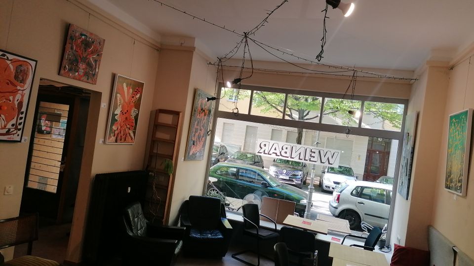 Innenraum eines kleinen Cafés oder einer Galerie mit mehreren Tischen und Stühlen sowie Sesselbereich, beige Wände sind mit großformatigen Gemälden dekoriert. Große Schaufensterfront zur Straße mit Blick auf parkende Autos; an der Decke verlaufen Lichterketten und Spots.