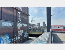 Moderne Stadtansicht mit einem mit Graffiti bedeckten Gebäude im Vordergrund, darüber Fenster mit der Aufschrift „WERK“. Im Hintergrund sind Bahngleise, eine Brücke, Hochhäuser und der Berliner Fernsehturm zu sehen. Rechts steht ein Rettungswagen.