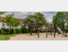 Ein Spielplatz mit Holzklettergerüst, Hängebrücken und einer Rutsche, umgeben von Bäumen und Sträuchern, im Hintergrund Reihenhäuser mit roten Ziegeldächern. Der Boden des Spielplatzes ist mit Sand bedeckt, daneben steht eine Parkbank auf einer Grünfläche.