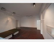 Ein minimalistisch gestalteter Raum mit weißen Wänden, Betonboden in rötlicher Farbe und einer schlichten Decke. Der Raum enthält eine eingelassene Sitzbank mit dunklen Polstern und ist mit einfachen Beleuchtungselementen sowie einer seitlichen Tür ausgestattet.