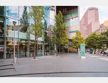 Außenansicht des Sony Centers in Berlin mit modernen Glasfassaden, Bäumen entlang eines gepflasterten Platzes und einem IMAX-Kino-Logo sichtbar. Im Hintergrund sind hohe, moderne Gebäude zu erkennen.