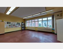 Klassenzimmer mit gelb gestrichenen Wänden, großen Fenstern auf der rechten Seite, die den Blick auf ein Gebäude und Bäume freigeben. An der linken Wand befindet sich eine Pinnwand mit Kinderzeichnungen sowie ein Regal mit Büromaterialien, und der Boden ist rötlich-braun.