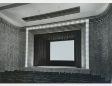 Schwarz-weiß-Fotografie eines historischen Kino- oder Theatersaals mit einer großen Bühne, auf der eine Leinwand hängt. Der Raum verfügt über gestufte, dekorative Wandverkleidungen und Sitzreihen im Vordergrund.