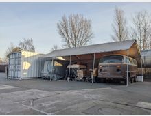 Ein überdachter Stellplatz mit einem alten, braunen VW-Bus und mehreren gelagerten Gegenständen, teils mit Planen abgedeckt. Rechts neben dem Stellplatz stehen Bäume, links ist ein weißer Frachtcontainer sichtbar.