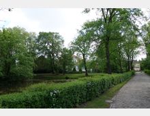 Parklandschaft mit einem kleinen Teich, umgeben von hohen, grünen Bäumen und gepflegten Hecken. Im Hintergrund sind ein weißes Gebäude mit roten Dachziegeln und ein gepflasterter Weg sichtbar.