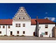 Weißes historisches Gebäude mit auffälligem verziertem Ziergiebel und roten Ziegeldächern. Das Gebäude hat mehrfach unterteilte Fenster und steht an einem gepflasterten Platz; rechts daneben befindet sich ein Informationsschild.