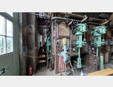 Industrieanlage mit Backsteinwänden und grünen, massiven Maschinenkomponenten, darunter Rohre und Ventile, die an einer Wand montiert sind. Im Vordergrund eine Tür mit Glaseinsätzen und ein Feuerlöscher; im Hintergrund eine Reihe von Stühlen und schmales Innenraumlicht.