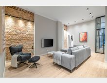 Modernes Wohnzimmer mit freigelegten Backsteinwänden, hellem Holzboden und deckenbündigen Spotleuchten. Ein großes graues Sofa und ein schwarzer Sessel mit Fußhocker stehen vor einer Wand mit Fernseher; daneben führt eine gläserne Schiebetür in einen angrenzenden Raum.