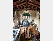 Innenansicht eines Gebäudes mit einem hohen, holzverkleideten Deckenbalken und Elementen im Stil einer umgestalteten Kapelle. Im Zentrum steht ein großer Holztisch mit Stühlen, dahinter eine Buddha-Statue vor einer halbrunden Nische mit buntem Mosaik und mehreren kleinen Fenstern.