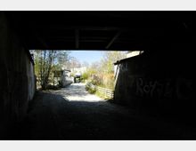 Ein gepflasterter Weg unter einer dunklen Brücke, der zu einem sonnigen Außenbereich mit einigen Gebäuden, Bäumen und einem geparkten Auto im Hintergrund führt. Die Brückenwände sind teils mit Graffiti versehen, und der Weg wird von einfachen Holzzäunen begrenzt.