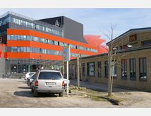 Ein moderner Bürokomplex mit einer auffälligen roten und schwarzen Fassade im Hintergrund, daneben ein niedriges Gebäude aus gelben Ziegeln mit großen Fenstern. Im Vordergrund parken Autos auf einem unbefestigten Platz, umgeben von jungen, unbelaubten Bäumen.