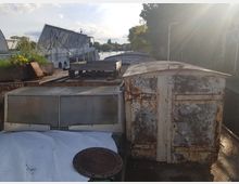 Ein alter, rostiger Schiffsaufbau mit Metalltüren und weiteren metallischen und hölzernen Gegenständen auf dem Deck, umgeben von Wasser mit Bäumen und Gebäuden im Hintergrund unter einem leicht bewölkten Himmel.