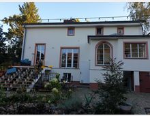 Zweistöckiges Wohnhaus mit heller Fassade und rot umrandeten Fenstern, flankiert von einem kleinen Garten mit Beeten, Pflanzentöpfen und einer Steintreppe. Im Vordergrund ist eine gepflasterte Fläche mit Pflanzen und einer kleinen Steinmauer zu sehen.