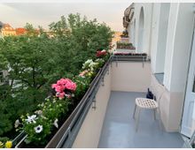 Ein schmaler Balkon mit hellgrauem Boden und weißen Wänden, geschmückt mit Blumenkästen voller bunter Blumen entlang des Geländers. Auf dem Balkon steht ein heller Kunststoffstuhl, und im Hintergrund sind grüne Baumwipfel sowie Gebäude mit roten Dächern sichtbar.