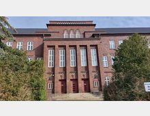 Backsteinfassade eines mehrstöckigen Gebäudes im Stil der Backsteinarchitektur mit hohen, rechteckigen Fenstern und großen, roten Türen im Erdgeschoss. Im Vordergrund befinden sich Büsche und ein Schild mit Hinweis auf eine Privatstraße.