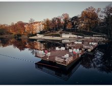 A serene waterfront scene with wooden floating platforms furnished with chairs and tables, surrounded by calm reflective water. In the background, there are trees in autumn colors, a hillside with pathways, and several white multi-story buildings nestled among the foliage.
