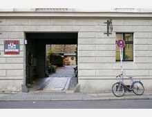 Ein heller Gebäudeeingang mit einem Durchgang zu einem gepflasterten Innenhof; daneben ein Schild mit der Aufschrift „Zur Alten Schmiede Lokal“. Im Vordergrund steht ein Fahrrad an einem Pfosten mit einem Halteverbotsschild.
