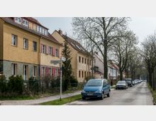 Straßenszene in einem Wohngebiet mit Reihenhäusern im Stil der frühen 20. Jahrhunderts; die Fassaden sind pastellgelb gestrichen, teilweise mit roten Fensterläden. Die Straße ist gesäumt von Bäumen mit beginnendem Blattwerk, geparkten Autos und einem Gehweg mit Kopfsteinpflaster.
