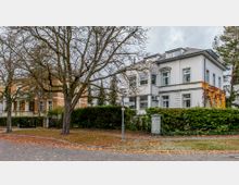 Ecke einer Wohnstraße mit zwei Villen: Links ein ziegelrotes Gebäude mit Säulen und Balkonen, rechts eine weiß verputzte Villa mit Efeubewuchs. Im Vordergrund ein kahler Baum, Laub auf dem Gehweg und ein Straßenschild mit den Namen „Hochkampstraße“ und „Marienstraße“.