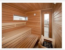 Kompakte Sauna aus Holz mit Bänken und einem Saunaofen im Vordergrund. Ein kleines Fenster sowie eine Glastür bieten Blick nach draußen.