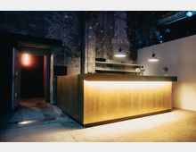 Moderner Barbereich in einem industriellen Interieur mit rohen, unverputzten Wänden. Der Tresen ist goldfarben beleuchtet, darüber hängen drei schwarze Hängeleuchten, und im Hintergrund sind Regale mit Gläsern erkennbar.