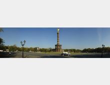 Panorama der Berliner Siegessäule auf dem Großen Stern, mit der goldenen Statue "Victoria" an der Spitze, umgeben von einer kreisförmigen Straße und Bäumen im Hintergrund. Im Vordergrund sind Laternenpfähle, eine vorbeifahrende schwarze und weiße Autos sowie Fußgänger sichtbar.