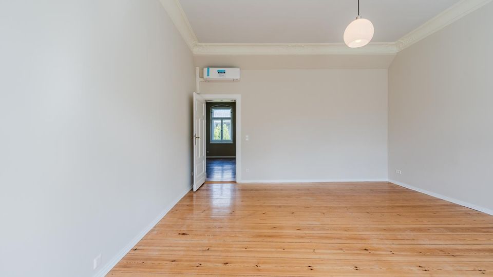 Leerer, heller Wohnraum mit hohen Decken, weißen Wänden und breitem Holzdielenboden. Eine runde Deckenleuchte hängt mittig, und durch eine offene Tür ist ein angrenzender Raum mit Fenster zu sehen.