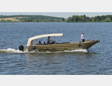 Ein offenes Motorboot mit einem beigen Sonnendach fährt auf einem weitläufigen See. Im Hintergrund sind Bäume, grüne Uferlandschaft und Windräder zu sehen.