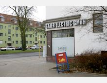 Straßenecke mit einem Ladenlokal, beschriftet mit „Saier Electric GmbH“, daneben ein Werbeschild für ein „Café“. Im Hintergrund sind parkende Autos, eine Straße und ein grünes Wohngebäude mit roten Dachziegeln zu sehen.