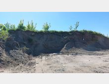 Eine freiliegende Erdböschung mit vereinzeltem Bewuchs an der Oberkante, darunter eine ebene, von Reifenspuren durchzogene Schotter- und Erdfläche, unter einem klaren blauen Himmel.