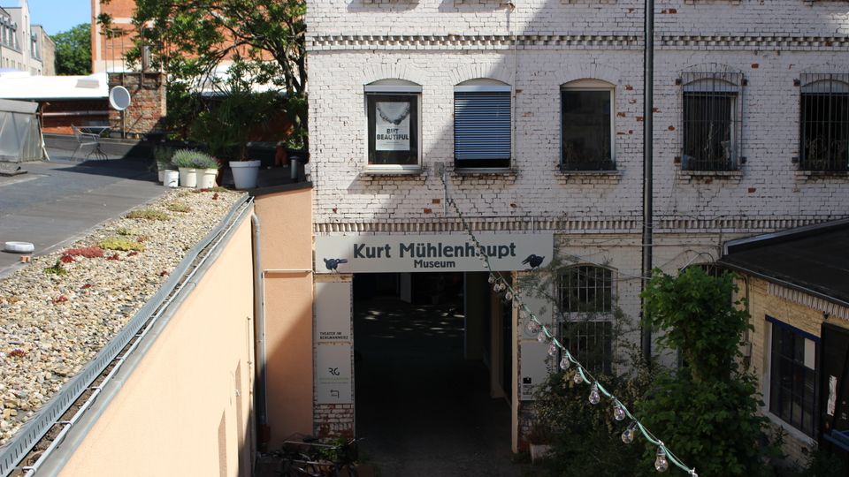 Eingang zu einem Hofdurchgang mit Schild „Kurt Mühlenhaupt Museum“ an einem weiß gestrichenen Backsteingebäude mit Rundbogenfenstern und teils herabgelassenen Rollläden. Links ist eine flache Dachfläche mit Kies und Pflanzenkübeln zu sehen, rechts begrenzen Bewuchs und eine Lichterkette den schmalen Zugang.