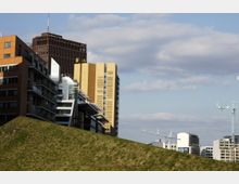 Moderne Stadtlandschaft mit mehreren Gebäuden, darunter ein auffälliger gelber Hochhauskomplex und ein dunkler Büroturm. Im Vordergrund ist eine grasbedeckte Anhöhe, während im Hintergrund Baustellenkräne und weitere urbane Strukturen zu sehen sind.
