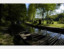 Ein ruhiger Wasserkanal mit aneinandergereihten Holzbooten entlang eines bewaldeten Ufers; rechts eine Wiese mit Sitzbänken, Bäumen und einem kleinen Pavillon im Sonnenschein.