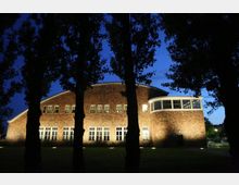 Backsteinbau mit halbrunder Fensterfront und mehreren rechteckigen Fenstern, beleuchtet in der Abenddämmerung, mit Bäumen im Vordergrund. Der Himmel ist tiefblau, und umliegende Lichter erhellen den Rasen.