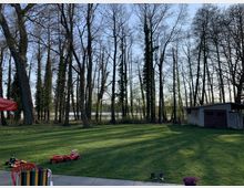 Ein Garten mit einer großen Rasenfläche, umgeben von hohen, kahlen Bäumen im Hintergrund, durch die ein Gewässer sichtbar ist. Rechts im Bild steht ein kleines Gartenhaus, und im Vordergrund befinden sich Spielzeug und ein bunter Stuhl auf einer Terrassenfläche.