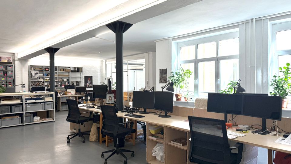 Heller, offener Büroraum im Loftstil mit langen Arbeitstischen, mehreren Monitoren und schwarzen Drehstühlen. Große Fensterfront mit Pflanzen auf der Fensterbank sowie offene Regale und Stauraumsysteme im Hintergrund; schwarze Stützsäulen tragen die Decke.