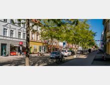 Straßenszene mit Bäumen, Sitzbänken und geparkten Autos entlang einer gepflasterten Gehwegfläche; links historische Gebäude mit verzierten Fassaden, darunter Geschäfte und Cafés. Über der Straße hängen bunte Dekorschirme, und Menschen spazieren oder verweilen im Schatten.