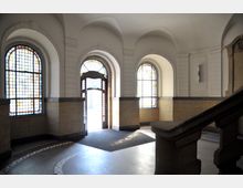 Eingangsbereich eines historischen Gebäudes mit hohen Rundbogenfenstern aus farbigem Glas, darunter ein offenes, verziertes Holztor, sowie einer geschwungenen Treppe rechts im Bild. Die Wände sind weiß gestrichen und mit gelblichen Kacheln im unteren Bereich verkleidet.