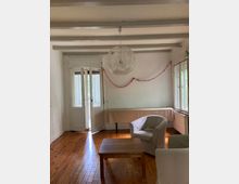 Wohnzimmer mit Holzboden, weißen Wänden und einer sichtbaren Holzbalkendecke. Im Raum stehen zwei Sessel und ein Holztisch, im Hintergrund ein langer Tisch mit Tischtuch vor einer Glas-Doppeltür, die auf einen Balkon oder in einen Garten führt. Eine runde Hängelampe und Girlanden an der Wand ergänzen die Einrichtung.