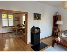 Blick in ein helles Wohn- und Esszimmer mit Holzfußboden: rechts ein schwarzer, runder Kaminofen vor weißer Wand neben Regal, Stehlampe und Sofa. Durch einen breiten Holzrahmen führt der Durchgang in den Essbereich mit rundem Holztisch, Stühlen, Pflanzen und großen Fenstern mit halb geschlossenen Plissees.