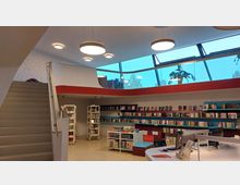 Eine moderne Bibliothek mit einer geschwungenen Treppe links, farbenfrohen Bücherregalen an der Wand und computergestützten Arbeitsplätzen im Vordergrund. Die Glasdecke im oberen Bereich lässt natürliches Licht herein und zeigt Pflanzen als Dekoration.